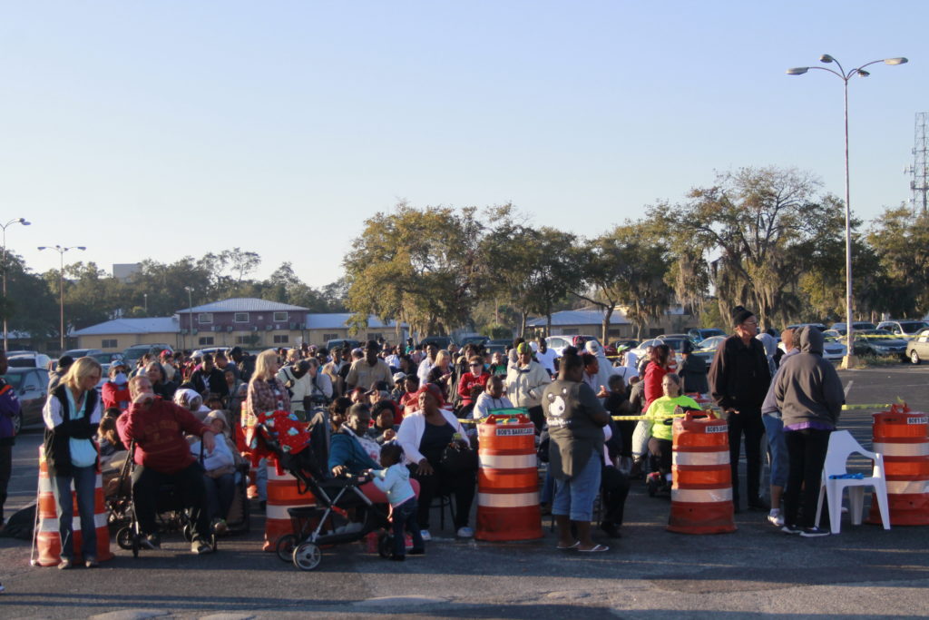 A large group of people, some with strollers, stand and sit in an outdoor area lined with orange traffic barrels. It's a sunny day with trees and parked cars in the background.