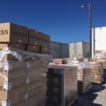 Stacks of cardboard boxes labeled "SNACKS" and other supplies are packed on wooden pallets in an outdoor storage area under clear blue Tampa Bay skies.