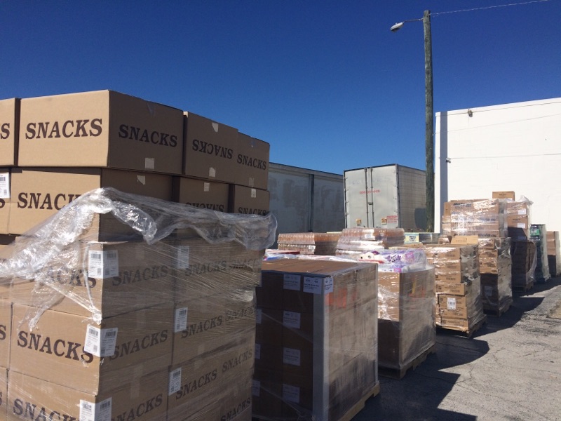 Stacks of cardboard boxes labeled "SNACKS" and other supplies are packed on wooden pallets in an outdoor storage area under clear blue Tampa Bay skies.