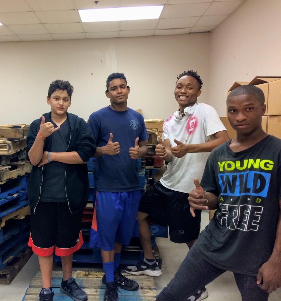 Four young men pose together in a room with wooden pallets, giving thumbs-up gestures and smiling at the camera. They are dressed casually in shorts and t-shirts, exuding a sense of camaraderie and shared purpose, possibly after participating in an event focused on charitable giving.