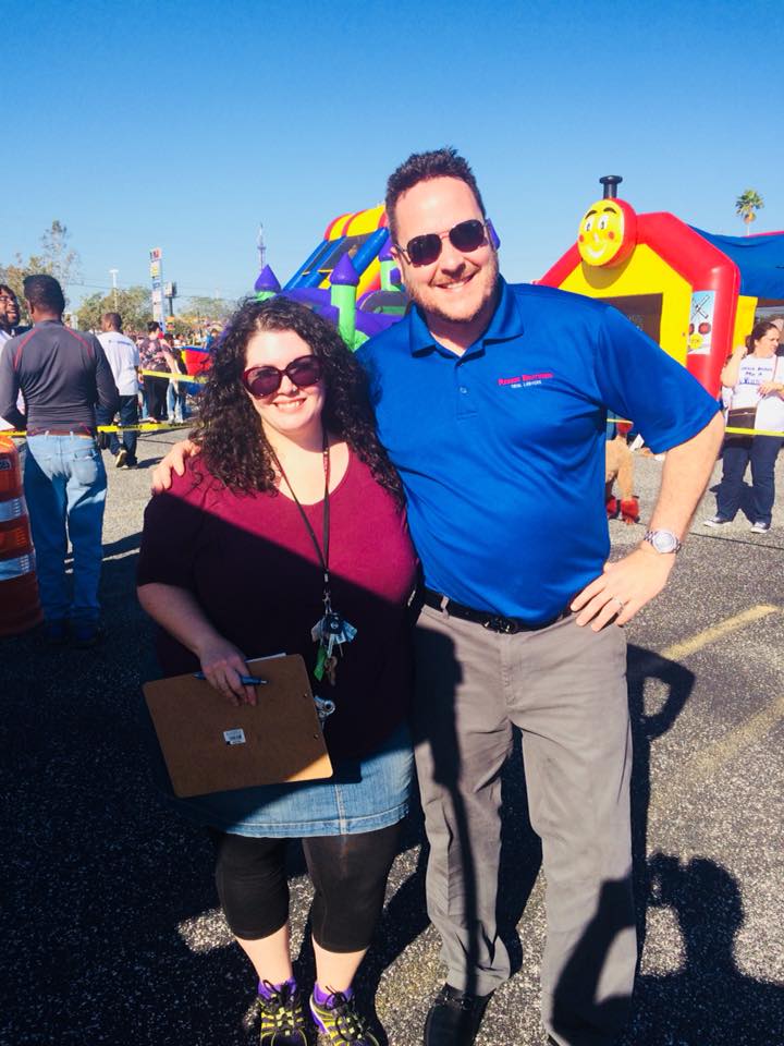 Two people pose together at an outdoor event with an inflatable play structure in the background. The person on the left, likely a volunteer, holds a clipboard, while the person on the right wears sunglasses and a blue shirt.