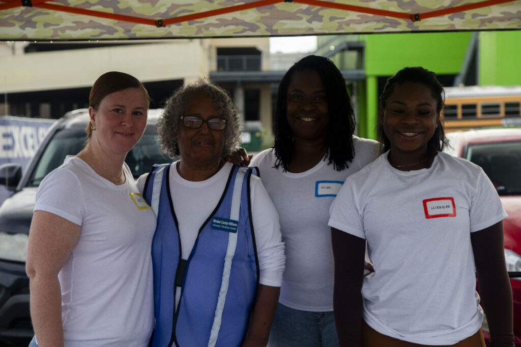 Four women, including Volunteer Josue, stand together under a canopy at an outdoor event, all smiling and wearing name tags, with cars and buildings visible in the background.
